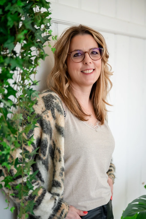 Christa Meier Therapeutin Christa Meier, Therapeutin für Bioresonanz, Hochfrequenz, Psych-k-coaching, langes, blondes Haar, Brille, freundlich lächelnd, graues Shirt, Jäckchen, halb hinter Efeu stehend